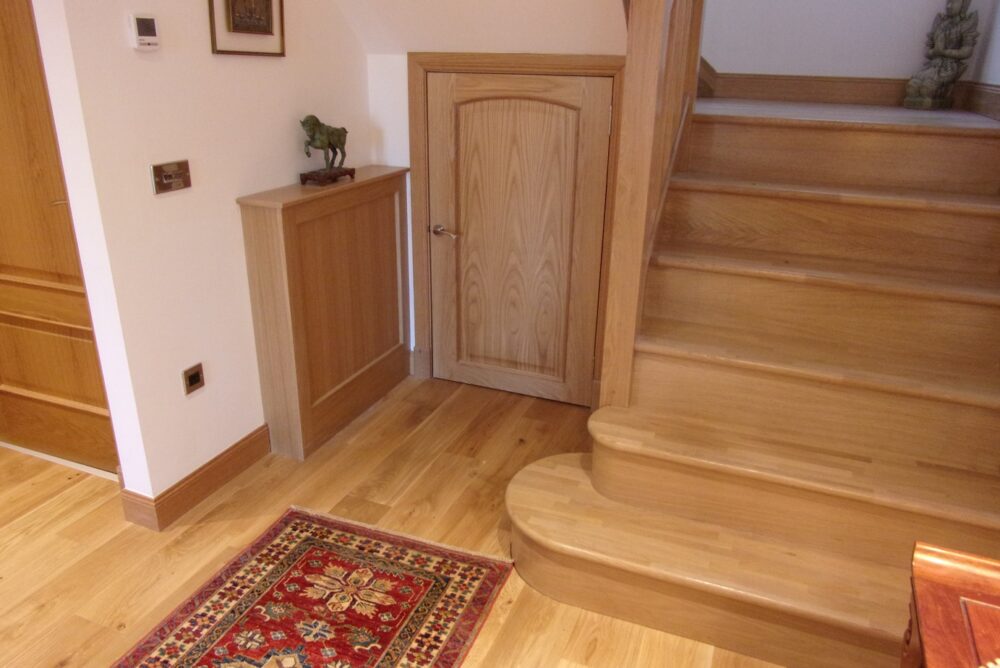 A small wooden door, crafted by a skilled Abernethy carpenter, is set beneath a staircase with wooden steps in a hallway with light wood floors and a patterned rug.