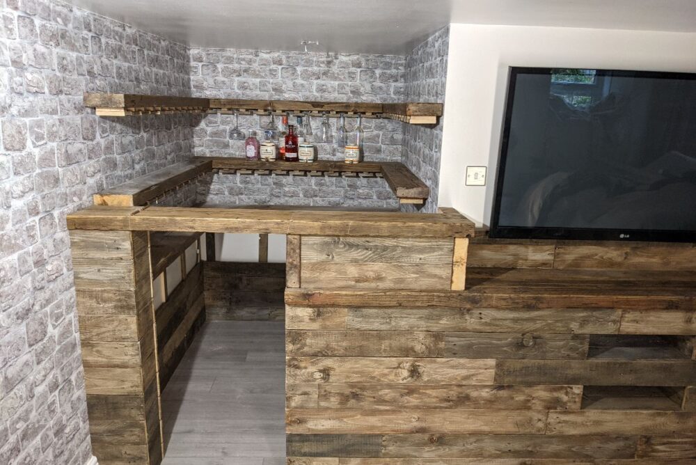 A rustic home bar with expert joinery, featuring a wood-paneled counter, shelves holding liquor bottles, and a wall-mounted TV on the right, set against brick-patterned wallpaper—perfectly crafted by a skilled Perth carpenter.
