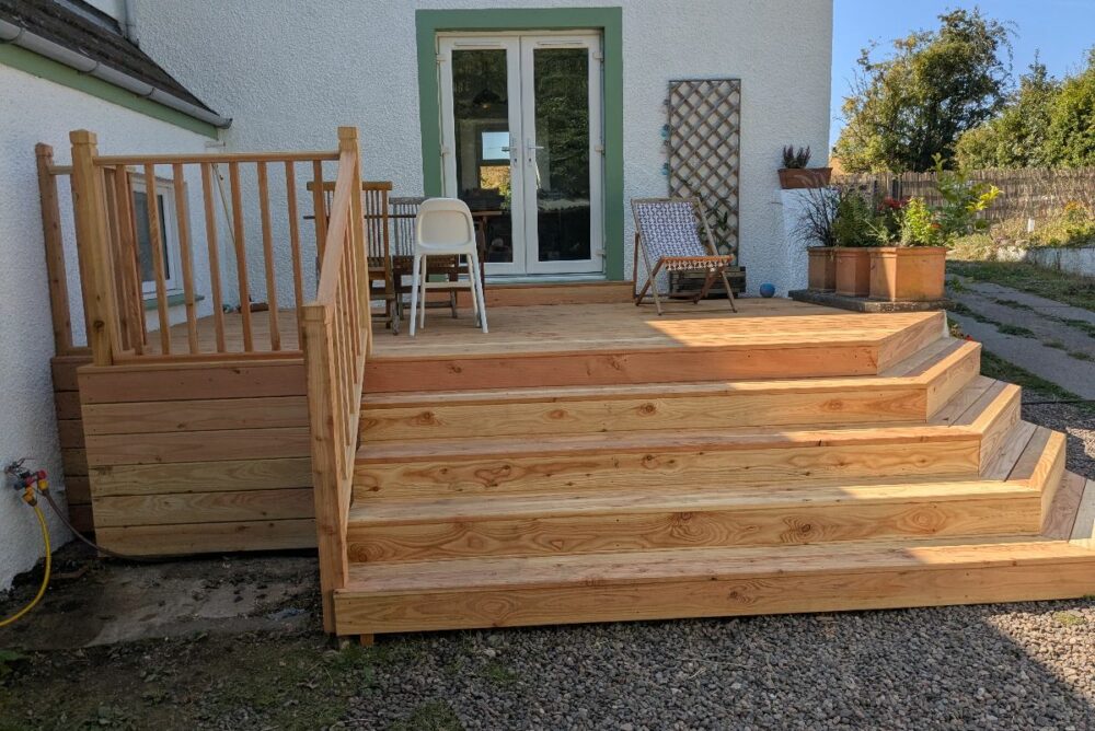 A wooden deck crafted by a skilled carpenter features steps leading to a patio area with chairs, a small table, and potted plants outside a white house with double glass doors in Abernethy, near Perth.