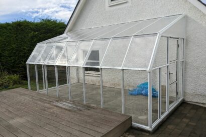 A glass greenhouse with a white frame stands on a tiled patio next to a white house in Abernethy, adjacent to a wooden deck and green hedge.