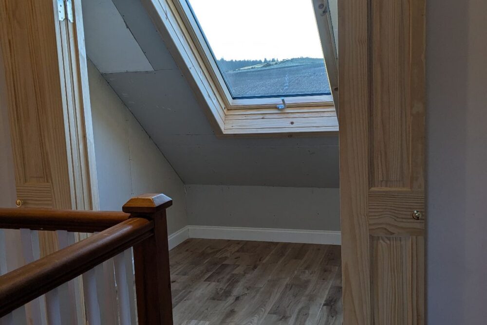 A small room with wood flooring, folding wooden doors crafted by a skilled carpenter, and a large skylight window on a sloped ceiling, overlooking an outdoor landscape near Abernethy.