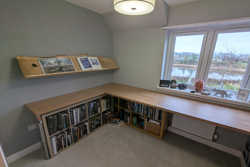 Corner home office in Perth with a wooden L-shaped desk crafted by a skilled carpenter; features a bookshelf under the desk, floating shelf with photos, and window overlooking Abernethy pond and trees.
