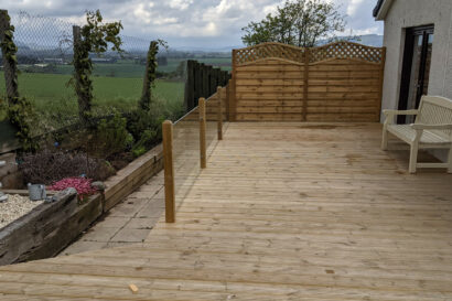 A wooden deck by Abernethy features glass panel railings, a wooden privacy screen crafted with expert joinery, a white bench, and a garden bed beside a fenced yard overlooking open fields.